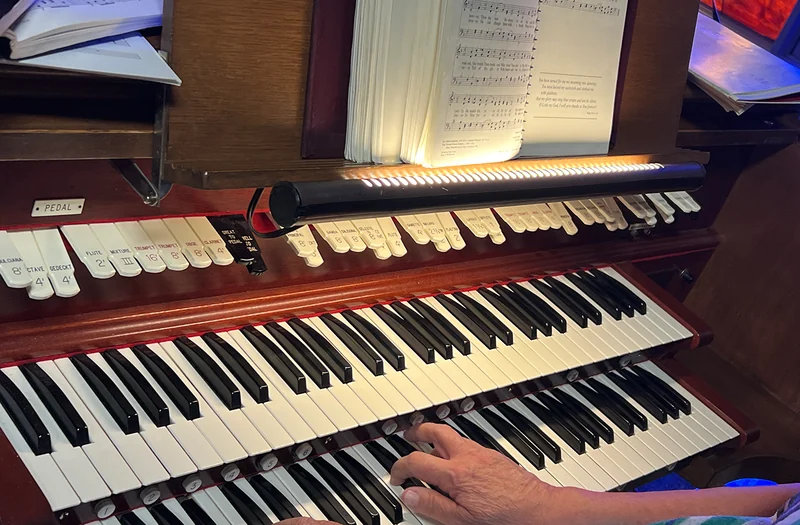 A hand playing Bethany’s wooden pipe-organ console, with its keyboards, stops and open music score.